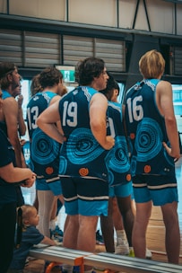 A group of young men wearing matching blue sports jerseys with tribal patterns stand indoors, likely in a gymnasium. The players are numbered, and their focus appears to be on something off-camera. A child sits on the bench nearby, adding an unexpected element to the scene.