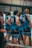 A group of young men wearing matching blue sports jerseys with tribal patterns stand indoors, likely in a gymnasium. The players are numbered, and their focus appears to be on something off-camera. A child sits on the bench nearby, adding an unexpected element to the scene.
