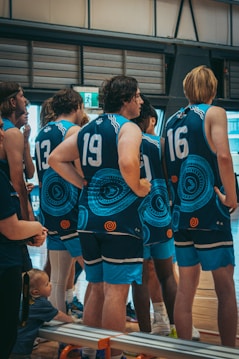 A group of young men wearing matching blue sports jerseys with tribal patterns stand indoors, likely in a gymnasium. The players are numbered, and their focus appears to be on something off-camera. A child sits on the bench nearby, adding an unexpected element to the scene.