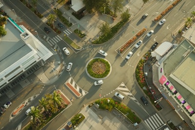 An aerial view of a roundabout surrounded by urban buildings and streets. Multiple vehicles navigate the circular traffic pattern, while landscaped greenery decorates the center and sides of the streets. The roads are well-marked with pedestrian crossings and traffic signage. The setting indicates a busy yet orderly urban environment with a mix of modern architecture and natural elements.