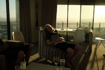 A person is reclining on an armchair in a dimly lit room, with sunlight streaming through large glass windows. The room contains modern furniture, including a table with white chairs. Outside, a balcony is visible with a view of distant mountains and a cityscape.
