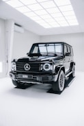 A black Mercedes-Benz G-Class SUV is positioned inside a modern, well-lit showroom. The vehicle's glossy finish reflects the overhead grid lights, and the room has a clean, minimalistic design with white walls and flooring.