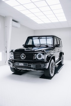 A black Mercedes-Benz G-Class SUV is positioned inside a modern, well-lit showroom. The vehicle's glossy finish reflects the overhead grid lights, and the room has a clean, minimalistic design with white walls and flooring.