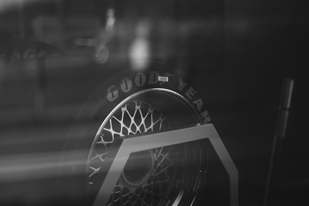 The black and white image features a close-up of a Goodyear tire. The tire appears to be on a display stand, with a reflective surface creating a somewhat abstract and moody ambiance. The tire's intricate tread pattern and the brand name are prominently visible.