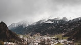 A small town nestled in a valley, surrounded by forested mountains with snow-capped peaks. The sky is overcast, casting a gray tone over the landscape. Buildings in the town appear small against the backdrop of the towering mountains.