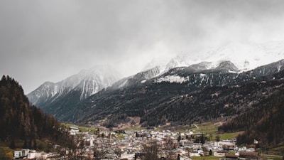 A small town nestled in a valley, surrounded by forested mountains with snow-capped peaks. The sky is overcast, casting a gray tone over the landscape. Buildings in the town appear small against the backdrop of the towering mountains.
