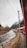 A red train travels alongside a winding river with a rocky shoreline. Power lines stretch above, and in the background, snow-capped mountains rise, partially shrouded by clouds. Sparse vegetation dots the landscape, giving a sense of early spring or autumn.