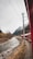 A red train travels alongside a winding river with a rocky shoreline. Power lines stretch above, and in the background, snow-capped mountains rise, partially shrouded by clouds. Sparse vegetation dots the landscape, giving a sense of early spring or autumn.