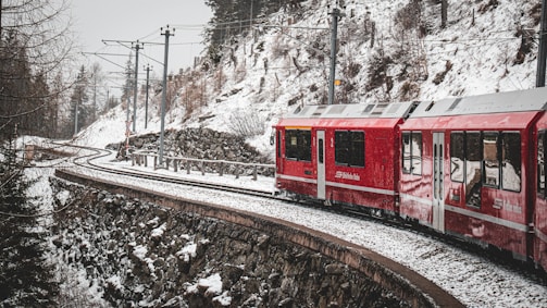 A red train is traveling through a snow-covered mountainous landscape. The scene features a curved railway track lined with overhead electrical lines, set against a backdrop of snow-dusted trees and rocky terrain.