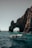 A glass-bottom boat navigates clear turquoise waters near a large natural rock arch formation. The sunlight casts shadows on the rugged surface of the rocks, and people in the boat are wearing life jackets.
