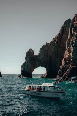 A glass-bottom boat navigates clear turquoise waters near a large natural rock arch formation. The sunlight casts shadows on the rugged surface of the rocks, and people in the boat are wearing life jackets.