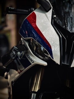 A close-up view of a motorcycle with sleek, sporty design features. The body has a glossy finish with prominent white, red, and blue colors. Metallic parts, such as the fork and handlebars, are visible, indicating a high-performance model.