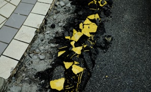 A cracked road surface with broken yellow paint scattered across the asphalt. The pavement on the left is composed of grey and white tiles, contrasting with the rough, dark texture of the road.