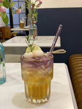 A vibrant cocktail sits elegantly on a marble table, featuring a blend of yellow and purple hues. The drink is garnished with a sprig of rosemary, a lime slice, and what appear to be lychee fruits. In the background, there are delicate pink flowers in vases, adding a touch of nature to the setting.