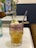 A vibrant cocktail sits elegantly on a marble table, featuring a blend of yellow and purple hues. The drink is garnished with a sprig of rosemary, a lime slice, and what appear to be lychee fruits. In the background, there are delicate pink flowers in vases, adding a touch of nature to the setting.