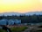 A row of modern residential houses is set against the backdrop of a forest and distant mountains at sunset. The sky transitions from yellow to blue as it meets the horizon. There is construction equipment in the foreground, indicating ongoing development.
