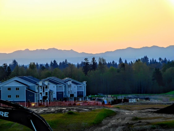 A row of modern residential houses is set against the backdrop of a forest and distant mountains at sunset. The sky transitions from yellow to blue as it meets the horizon. There is construction equipment in the foreground, indicating ongoing development.