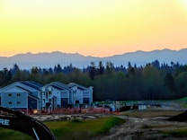 A row of modern residential houses is set against the backdrop of a forest and distant mountains at sunset. The sky transitions from yellow to blue as it meets the horizon. There is construction equipment in the foreground, indicating ongoing development.