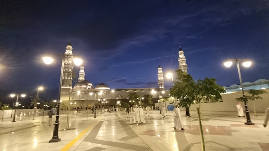 A grand mosque with multiple minarets is illuminated against a deep blue evening sky. Tall street lamps line the paved plaza, casting a warm glow. Trees are interspersed among the open space, and people dressed in traditional attire are walking around, contributing to a lively ambiance.