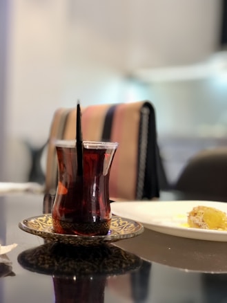A glass of dark-colored tea is served on an ornate saucer on a reflective table, with a piece of cake on a white plate in the background. The setting has a cozy and relaxed ambiance with soft lighting.