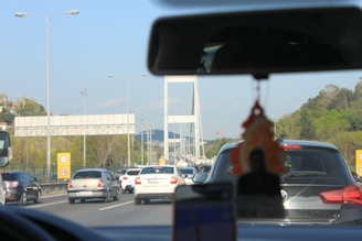 A crowded highway scene with numerous vehicles, including cars and vans, moving towards a large white suspension bridge in the background. The foreground contains the interior of a vehicle, showing a rear-view mirror and air freshener hanging with a blurred image. Trees and green foliage line the road on both sides, and overhead lampposts and road signs are visible.