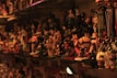 A densely packed shelf filled with a variety of anime and cartoon figurines, including keychains labeled with 'Naruto'. The lighting casts warm tones over the collection, creating a nostalgic and cluttered atmosphere. Various characters in bright and bold colors are displayed, with some recognizable to anime enthusiasts.
