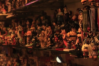 A densely packed shelf filled with a variety of anime and cartoon figurines, including keychains labeled with 'Naruto'. The lighting casts warm tones over the collection, creating a nostalgic and cluttered atmosphere. Various characters in bright and bold colors are displayed, with some recognizable to anime enthusiasts.