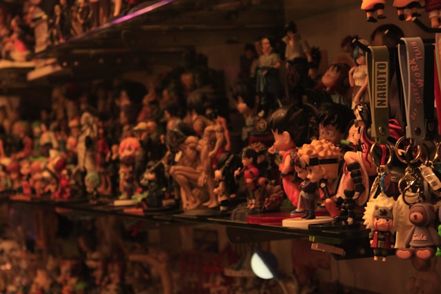 A densely packed shelf filled with a variety of anime and cartoon figurines, including keychains labeled with 'Naruto'. The lighting casts warm tones over the collection, creating a nostalgic and cluttered atmosphere. Various characters in bright and bold colors are displayed, with some recognizable to anime enthusiasts.