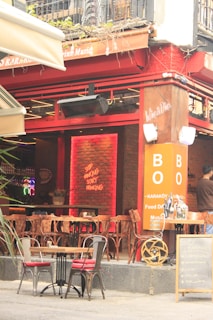 A cozy caf&eacute; with outdoor seating featuring wooden chairs and small tables adorned with red cushions. The interior has a warm, inviting atmosphere with a brick wall illuminated by red lighting and a sign reading 'Stop thinking, start drinking'. The exterior has orange signage with text and a small chalkboard menu. A person is seen standing at the entrance, adding to the lively ambiance.