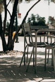 A set of outdoor chairs and a table is situated on a shaded patio. The metal furniture features a modern design and is surrounded by tree trunks and foliage. Sunlight creates dappled shadows on the ground, enhancing the tranquil atmosphere.