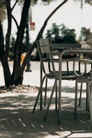 A set of outdoor chairs and a table is situated on a shaded patio. The metal furniture features a modern design and is surrounded by tree trunks and foliage. Sunlight creates dappled shadows on the ground, enhancing the tranquil atmosphere.