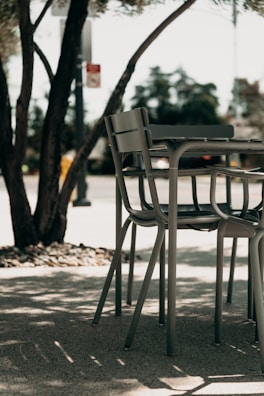 A set of outdoor chairs and a table is situated on a shaded patio. The metal furniture features a modern design and is surrounded by tree trunks and foliage. Sunlight creates dappled shadows on the ground, enhancing the tranquil atmosphere.