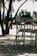 A set of outdoor chairs and a table is situated on a shaded patio. The metal furniture features a modern design and is surrounded by tree trunks and foliage. Sunlight creates dappled shadows on the ground, enhancing the tranquil atmosphere.