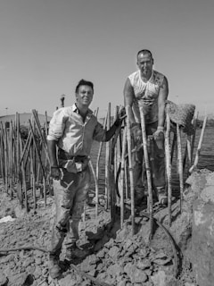 Two men are working in a construction-like setting near water. They are surrounded by concrete and rebar structures. One man is standing on rocky ground, while the other is positioned above him, resting his arms on the rebar.