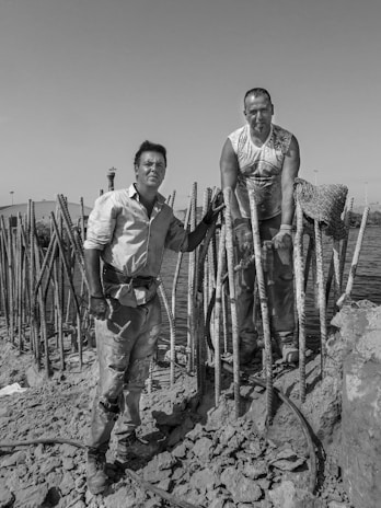 Two men are working in a construction-like setting near water. They are surrounded by concrete and rebar structures. One man is standing on rocky ground, while the other is positioned above him, resting his arms on the rebar.