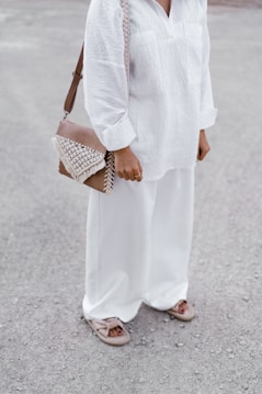 A person dressed in a white, loose-fitting shirt and wide-legged pants, with a woven leather handbag featuring fringe details hanging from the shoulder. They are wearing open-toed sandals and are standing on a gravel-like surface.