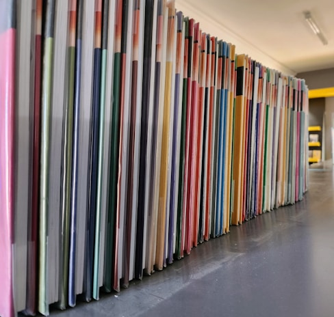 A neatly arranged row of colorful books or folders is lined up on a dark surface, with each cover adding a different hue to the sequence. The angle of the photo captures the long stretch of the arrangement, providing a sense of order and organization.
