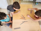 Three children are gathered around a wooden table, focusing intently on playing with a bundle of colored sticks laid out in front of them. The setting appears to be indoors, with a tiled floor and a variety of objects in the background, including a small soccer ball.