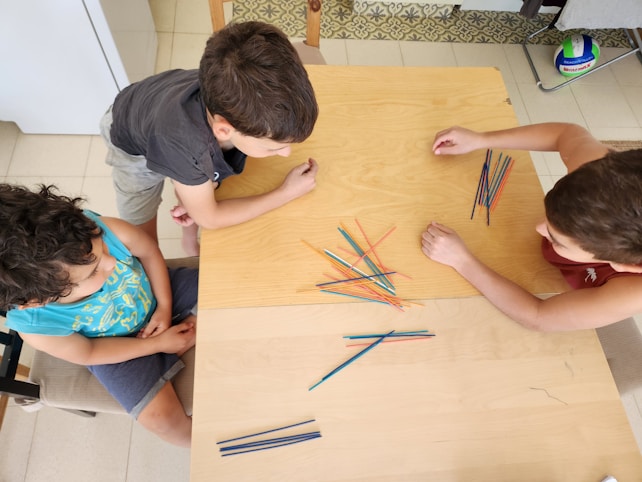 Three children are gathered around a wooden table, focusing intently on playing with a bundle of colored sticks laid out in front of them. The setting appears to be indoors, with a tiled floor and a variety of objects in the background, including a small soccer ball.