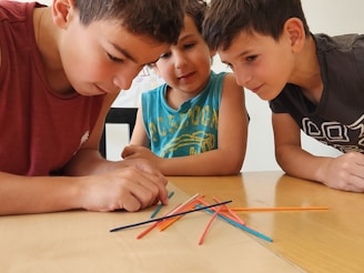 Three boys are deeply focused on what appears to be a game involving colorful wooden sticks, placed on a wooden table. They are leaning in closely, observing the sticks intently.