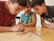 Three boys are deeply focused on what appears to be a game involving colorful wooden sticks, placed on a wooden table. They are leaning in closely, observing the sticks intently.