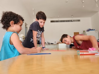 Three children are gathered around a wooden table in a bright, minimalist room. They appear to be engaged in a craft activity, with colored pencils and paper on the table. The background shows a kitchen area with a refrigerator and some supplies.