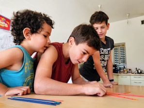 Three children are gathered around a table, intently focused on a colorful activity involving thin, colored sticks. The children appear to be in a classroom or home setting, with kitchen items and writing on a whiteboard visible in the background.
