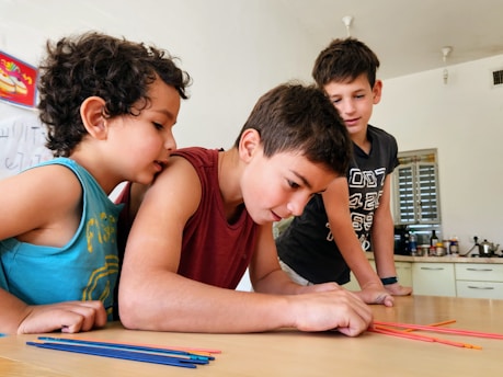 Three children are gathered around a table, intently focused on a colorful activity involving thin, colored sticks. The children appear to be in a classroom or home setting, with kitchen items and writing on a whiteboard visible in the background.