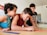 Three children are gathered around a table, intently focused on a colorful activity involving thin, colored sticks. The children appear to be in a classroom or home setting, with kitchen items and writing on a whiteboard visible in the background.