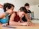 Three children are gathered around a table, intently focused on a colorful activity involving thin, colored sticks. The children appear to be in a classroom or home setting, with kitchen items and writing on a whiteboard visible in the background.