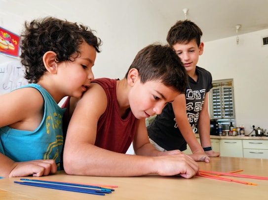 Three children are gathered around a table, intently focused on a colorful activity involving thin, colored sticks. The children appear to be in a classroom or home setting, with kitchen items and writing on a whiteboard visible in the background.