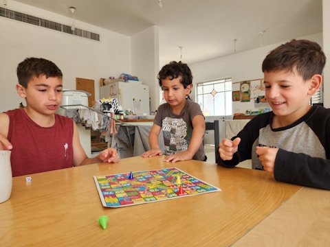Three children are gathered around a table playing a colorful board game. They seem engaged and focused on the game. The room appears to be a living space with various items like a fridge, clothes, and children's drawings visible in the background.