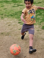 A young child is energetically playing soccer on a grassy field. The child is dressed in a beige t-shirt with a cartoon graphic and gray shorts, and is in mid-motion about to kick an orange soccer ball. The grass is a mix of green and brown patches, indicating a natural outdoor setting.