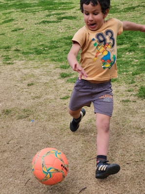 A young child is energetically playing soccer on a grassy field. The child is dressed in a beige t-shirt with a cartoon graphic and gray shorts, and is in mid-motion about to kick an orange soccer ball. The grass is a mix of green and brown patches, indicating a natural outdoor setting.